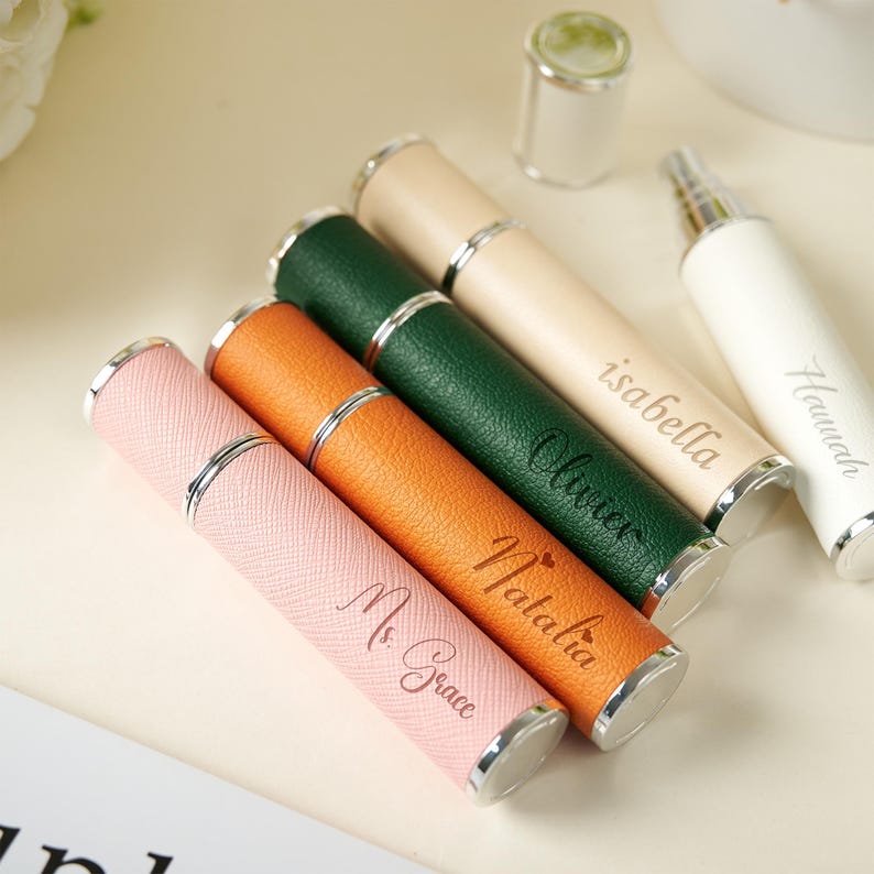 May include: A collection of personalized perfume atomizers in various colors, including pink, orange, green, beige, and white. Each atomizer has a silver-colored band and cap, with names engraved on the side. The atomizers are cylindrical in shape.