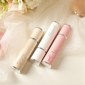May include: Three perfume atomizers in beige, white, and pink, each with a silver cap and base. The beige atomizer is labeled "Isabella", the white one "Hannah", and the pink one "Ms. Grace". The atomizers are cylindrical and appear to be made of leather.