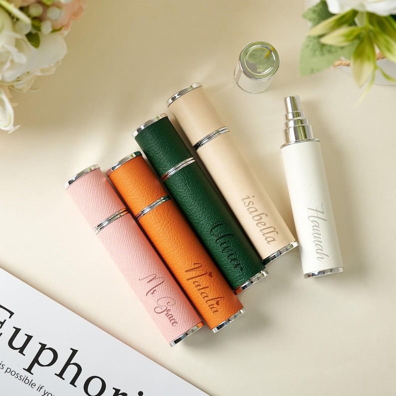 May include: A collection of personalized perfume atomizers in various colors, including pink, orange, green, and cream. Each atomizer has a silver-colored band and is engraved with a name. A white atomizer is also present.