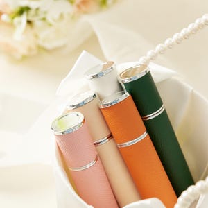 May include: A collection of colorful perfume atomizers in a white basket. The atomizers are in shades of pink, white, orange, and green, with silver accents. The background includes a pearl necklace and soft floral arrangements.