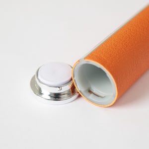 May include: An open, orange leather-wrapped tube with a silver cap and a white, circular pad. The tube's interior is white. The cap is detached, revealing the pad. The leather has a textured appearance.