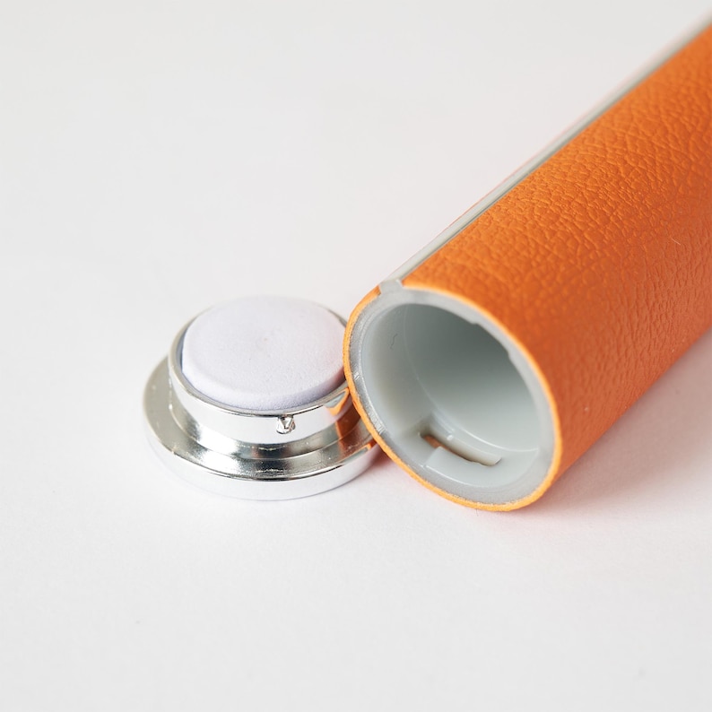 May include: An open, orange leather-wrapped tube with a silver cap and a white, circular pad. The tube's interior is white. The cap is detached, revealing the pad. The leather has a textured appearance.