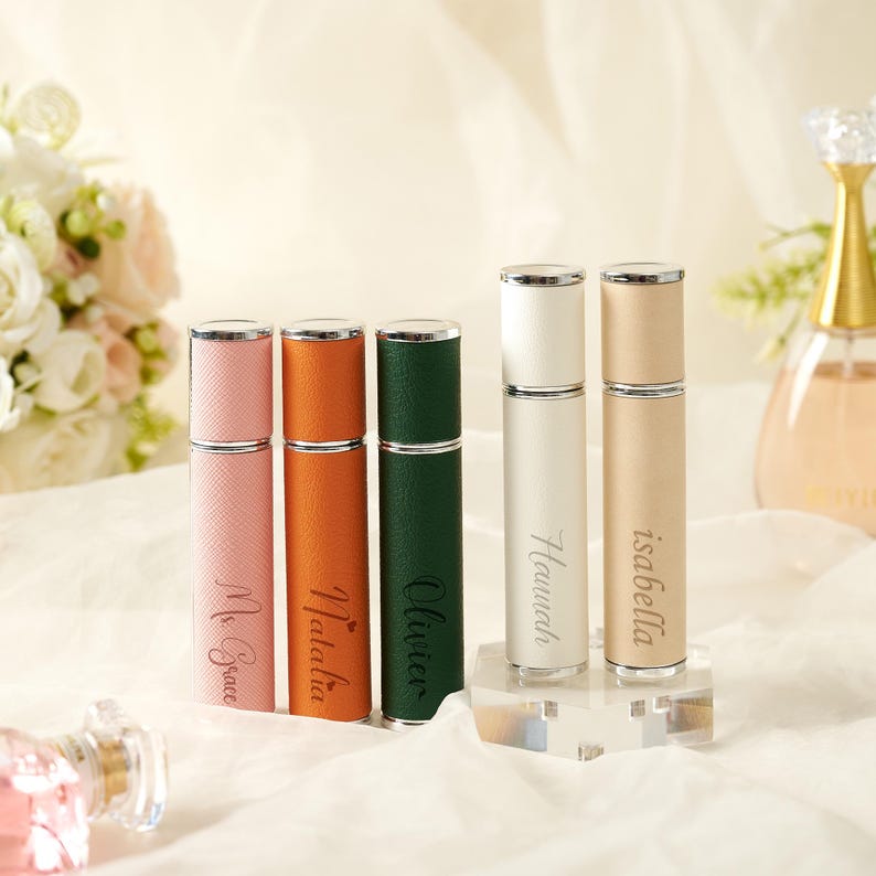 May include: A collection of personalized perfume atomizers in various colors, including pink, orange, green, white, and beige. Each atomizer has a silver cap and is engraved with a name in a cursive font. The atomizers are displayed on a clear stand.