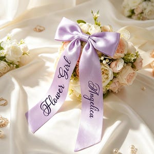 May include: A lavender satin ribbon bow featuring two long tails with black script text. The left tail displays the words Flower Girl and the right tail displays the name Angelica. The bow is positioned atop a cluster of cream and peach artificial roses.