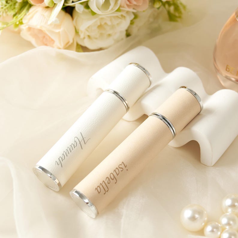 May include: Two personalized perfume atomizers. One is white with "Hannah" engraved, the other is beige with "isabella" engraved. Both have silver accents and are cylindrical. The atomizers rest on white pedestals, with flowers and pearls in the background.