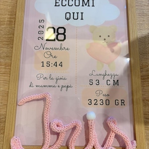 Op de afbeelding: Een ingelijste geboorteaankondiging met de naam "Iris" in roze garen. De aankondiging bevat de datum, tijd, lengte (53 cm) en gewicht (3230 g) van de baby, samen met de tekst "EccoMi Qui".