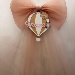 Pode incluir: Um grande laço de tule cor de pêssego com um design decorativo de balão de ar quente. O balão é branco e castanho com o nome "Ginevra" em escrita rosa. Uma pequena etiqueta de madeira diz "Eccomi" e um gráfico de ursinho de pelúcia.