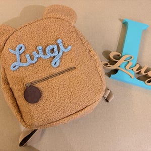 Op de afbeelding: Een beige rugzak in de vorm van een teddybeer van pluizige stof, met de naam Luigi in lichtblauwe schrijfletters op de voorkant, een ritsvak met een ronde bruine hanger en een grote turquoise letter L met de naam Luigi in hout.