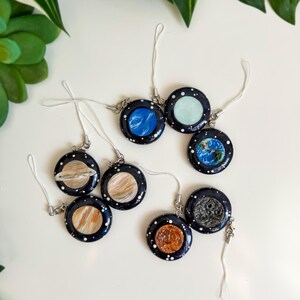 May include: A selection of circular charms, each representing a planet, with a dark blue background and white star details. The charms are attached to white cords. Planets such as Earth, Jupiter, and Saturn are visible, offering a celestial theme.