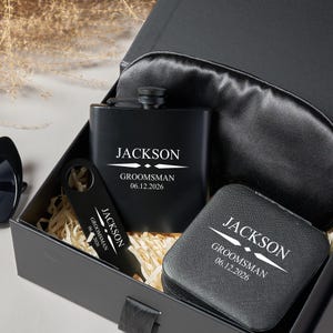 Peut inclure: Coffret cadeau noir contenant une flasque, un ouvre-bouteille, un masque de sommeil et un petit &eacute;tui. Chaque article est personnalis&eacute; avec le nom "JACKSON" et le texte "GROOMSMAN 06.12.2026" en blanc. La bo&icirc;te est remplie de paille d&eacute;corative.