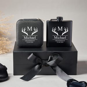 Peut inclure: Un coffret cadeau noir comprenant une flasque personnalis&eacute;e et un petit &eacute;tui, tous deux avec du texte blanc et un motif de bois de cerf. Le texte comprend la lettre "M", le nom "Michael" et la date "22.08.2026". L'ensemble est pr&eacute;sent&eacute; sur une bo&icirc;te noire avec un n&oelig;ud.