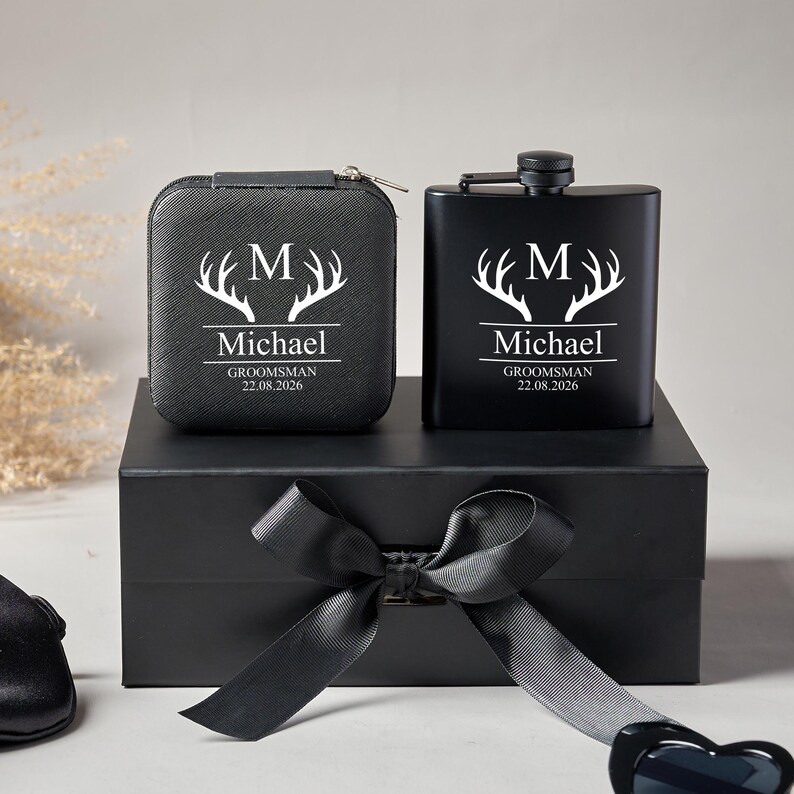 Peut inclure: Un coffret cadeau noir comprenant une flasque personnalis&eacute;e et un petit &eacute;tui, tous deux avec du texte blanc et un motif de bois de cerf. Le texte comprend la lettre "M", le nom "Michael" et la date "22.08.2026". L'ensemble est pr&eacute;sent&eacute; sur une bo&icirc;te noire avec un n&oelig;ud.