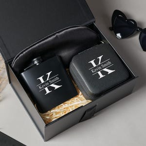 Peut inclure: Un coffret cadeau noir contenant une flasque, un petit &eacute;tui et un masque de sommeil. La flasque et l'&eacute;tui sont noirs avec les initiales "K" et le nom "Kevin Smith" en blanc. Le masque de sommeil est noir. Des lunettes de soleil en forme de c&oelig;ur sont visibles.