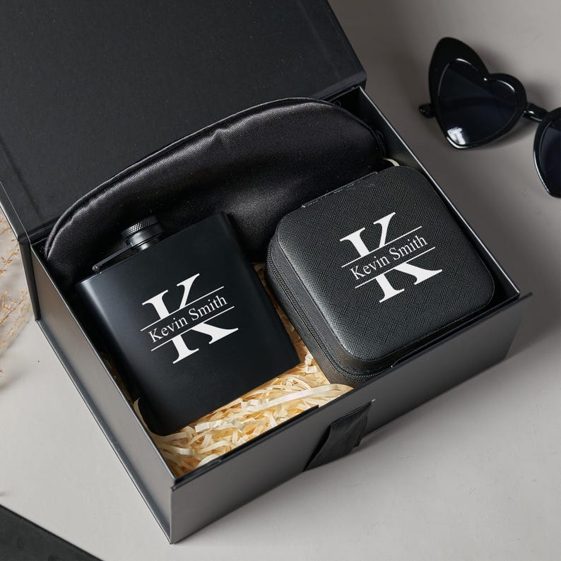 Peut inclure: Un coffret cadeau noir contenant une flasque, un petit &eacute;tui et un masque de sommeil. La flasque et l'&eacute;tui sont noirs avec les initiales "K" et le nom "Kevin Smith" en blanc. Le masque de sommeil est noir. Des lunettes de soleil en forme de c&oelig;ur sont visibles.