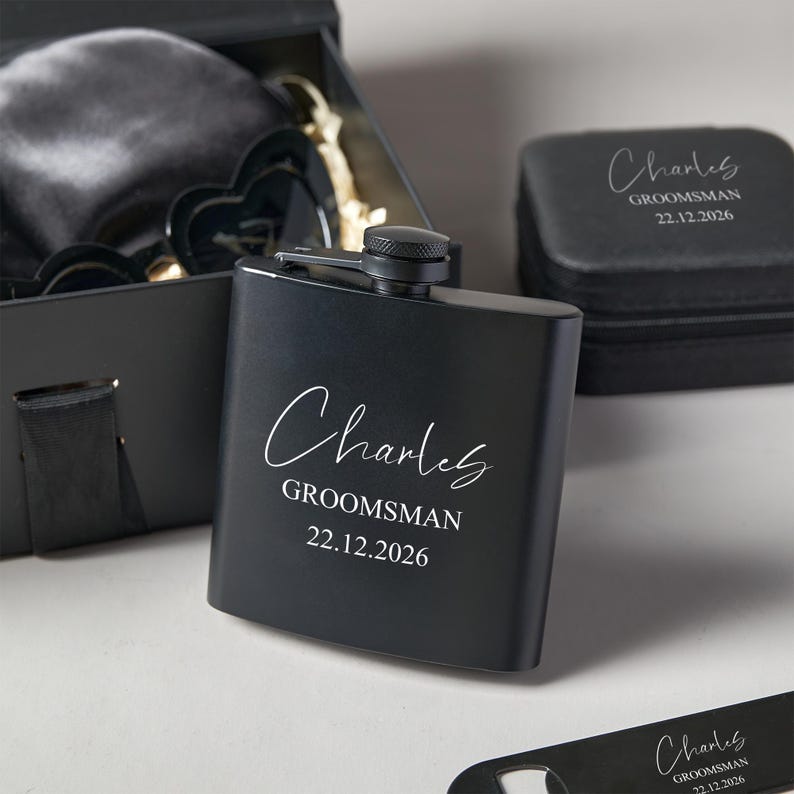Peut inclure: Une flasque noire personnalis&eacute;e avec le nom "Charles", le mot "Groomsman" et la date "22.12.2026" en &eacute;criture blanche. La flasque fait partie d'un coffret cadeau, comprenant une bo&icirc;te noire et d'autres accessoires. Id&eacute;al pour un mariage.