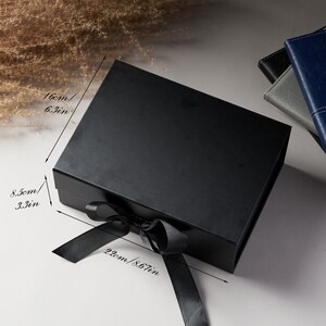 Peut inclure: Une bo&icirc;te cadeau noire avec un n&oelig;ud en ruban de satin. La bo&icirc;te mesure 16 cm sur 22 cm et fait 8,5 cm de haut. La bo&icirc;te est ferm&eacute;e et repose sur une surface blanche. D'autres carnets sont visibles en arri&egrave;re-plan.