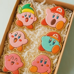 May include: A box of decorated sugar cookies featuring the character Kirby. The cookies are pink, orange, and blue, with various expressions and hats. The cookies are in a brown box with paper filler.