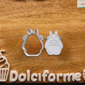 Puede incluir: Cortadores de galletas de pl&aacute;stico blanco con forma de cupcake, un personaje de Totoro y un contorno con forma de Totoro. La palabra "Dolciforme" est&aacute; escrita con letras blancas. El fondo es una superficie de madera.