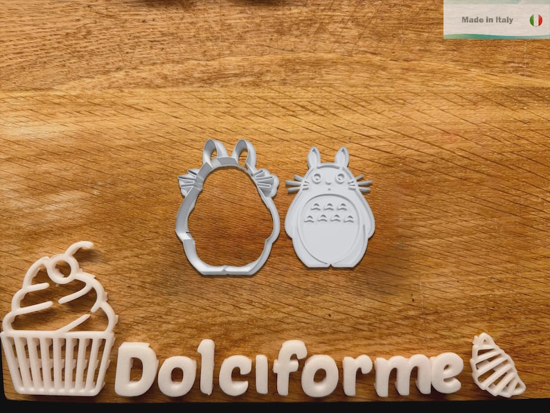Puede incluir: Cortadores de galletas de pl&aacute;stico blanco con forma de cupcake, un personaje de Totoro y un contorno con forma de Totoro. La palabra "Dolciforme" est&aacute; escrita con letras blancas. El fondo es una superficie de madera.