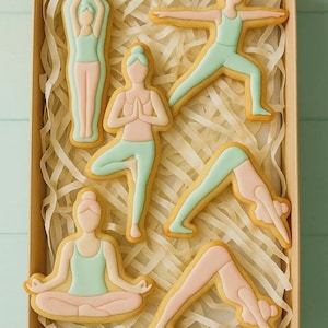 May include: A box of yoga-themed cookies. Each cookie is shaped like a person in a yoga pose, with light pink and mint green icing. The cookies are arranged on a bed of shredded paper inside a brown box.