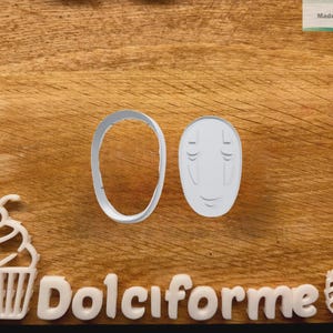 Puede incluir: Cortadores de galletas de pl&aacute;stico blanco con forma de cara y &oacute;valo, con la palabra "Dolciforme" en letras blancas. Tambi&eacute;n hay una magdalena y un cortador en forma de concha. El fondo es una superficie de madera.
