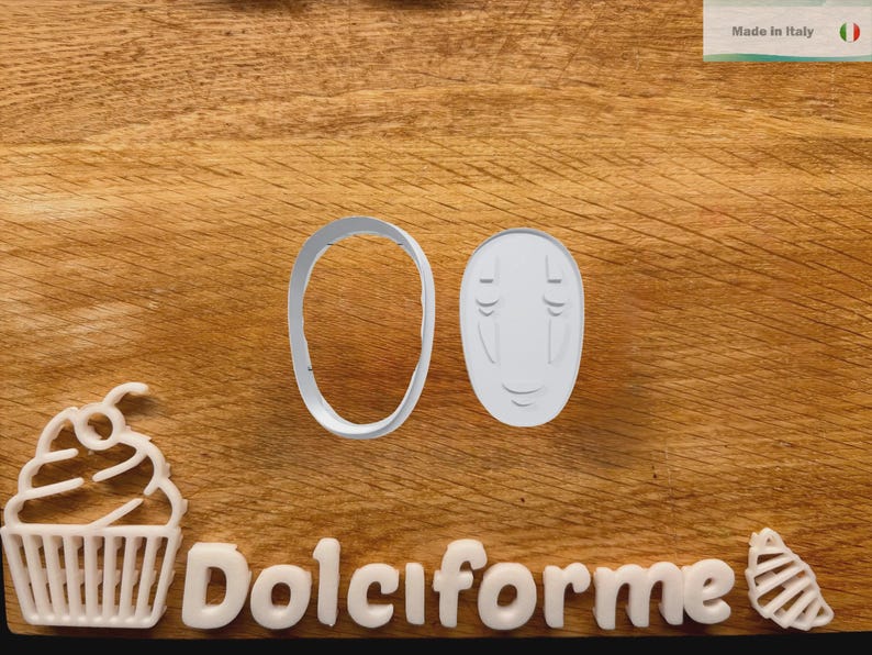 Puede incluir: Cortadores de galletas de pl&aacute;stico blanco con forma de cara y &oacute;valo, con la palabra "Dolciforme" en letras blancas. Tambi&eacute;n hay una magdalena y un cortador en forma de concha. El fondo es una superficie de madera.