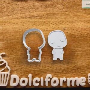Puede incluir: Dos cortadores de galletas blancos con forma de personaje de dibujos animados, uno s&oacute;lido y otro con contorno. Tambi&eacute;n se ven una magdalena blanca y la palabra "Dolciforme". El fondo es una superficie de madera.