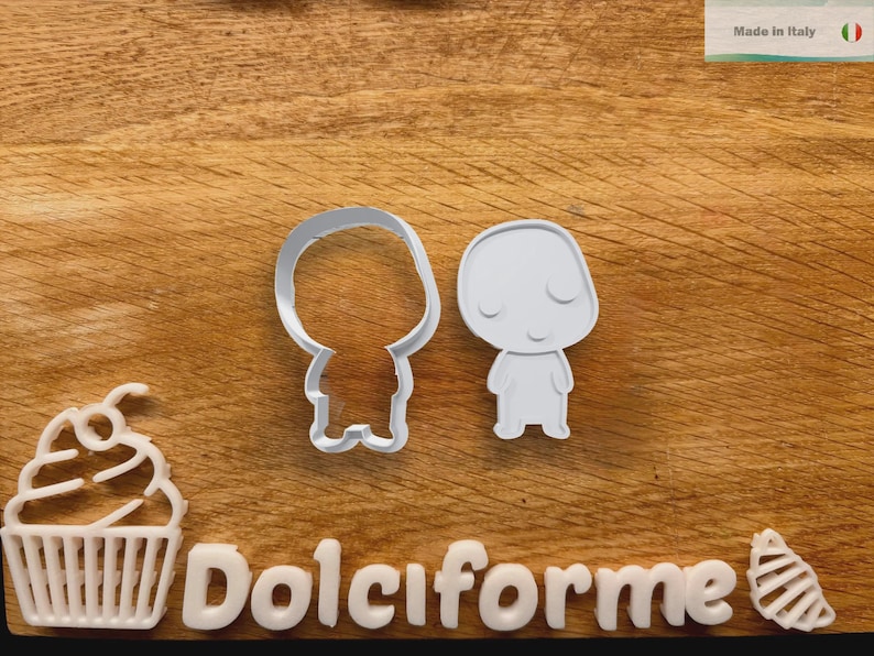 Puede incluir: Dos cortadores de galletas blancos con forma de personaje de dibujos animados, uno s&oacute;lido y otro con contorno. Tambi&eacute;n se ven una magdalena blanca y la palabra "Dolciforme". El fondo es una superficie de madera.