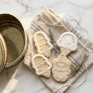 Op de afbeelding: Vier suikerkoekjes versierd met wit glazuur. Twee koekjes hebben de vorm van ijshoorntjes, één is een ijshoorntje met één bolletje en één is een teddybeer met een feesthoedje. De koekjes zijn gerangschikt op een beige linnen doek.