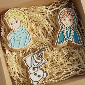 Puede incluir: Una caja con tres galletas decoradas que representan personajes de la película "Frozen". Las galletas, que representan a Elsa, Anna y Olaf, están decoradas con glaseado azul, blanco y marrón. Se encuentran sobre papel triturado.