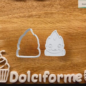 Puede incluir: Cortadores de galletas de pl&aacute;stico blanco con forma de cupcake, un personaje en forma de llama y una llama estilizada. La palabra "Dolciforme" est&aacute; escrita con letras blancas. El fondo es una superficie de madera.