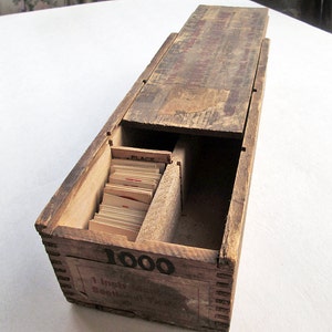 May include: A vintage wooden card catalog box with a sliding lid. The box is stained a dark brown and has visible wear. The front of the box reads "1000" and "1 inch Sectional Files". Inside are several small cards.