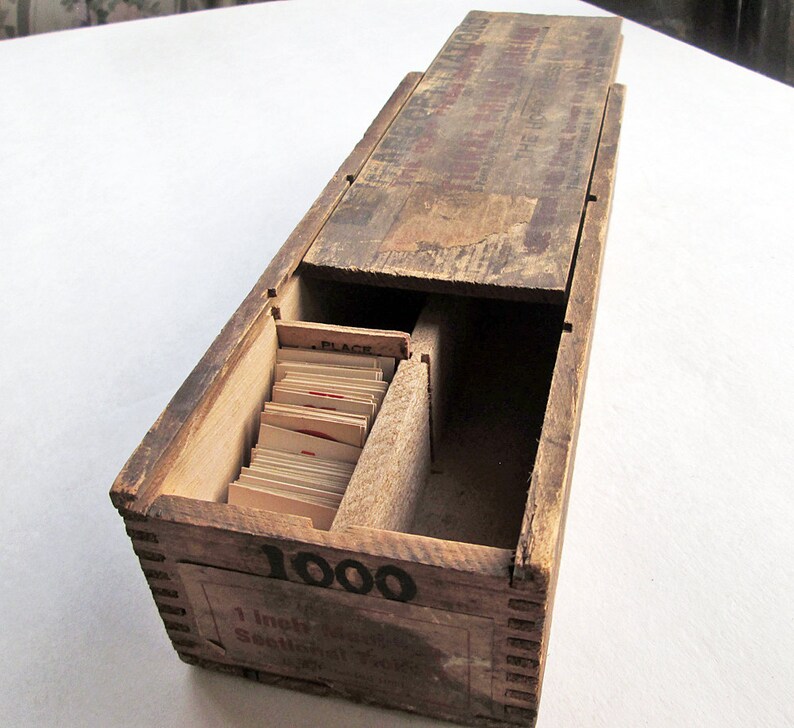 May include: A vintage wooden card catalog box with a sliding lid. The box is stained a dark brown and has visible wear. The front of the box reads "1000" and "1 inch Sectional Files". Inside are several small cards.