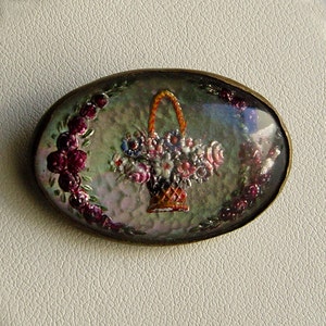 May include: Oval-shaped brooch with a floral basket design. The brooch features a basket filled with colorful flowers, surrounded by a wreath of red flowers and green leaves. The brooch has a gold-colored border.