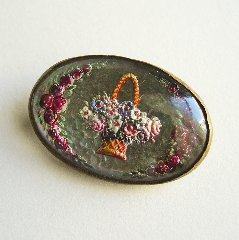 May include: Oval-shaped brooch with a floral design. The brooch features a basket filled with colorful flowers, surrounded by a wreath of red roses. The brooch has a gold-colored metal frame and a dark background.