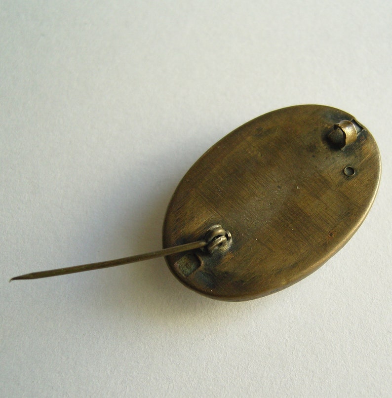 May include: An antique oval brooch with a pin closure. The brooch has a bronze-colored metal finish and a simple design. The pin is long and slender, and the clasp is a simple, functional design. The brooch is likely vintage.