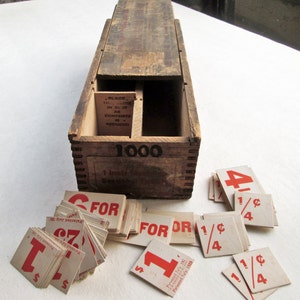 May include: A vintage wooden box with the number "1000" printed on the front, containing small square cards with red numbers and text. The cards read "1", "4", "1/4", and "FOR". The box has a sliding lid and a slot inside.