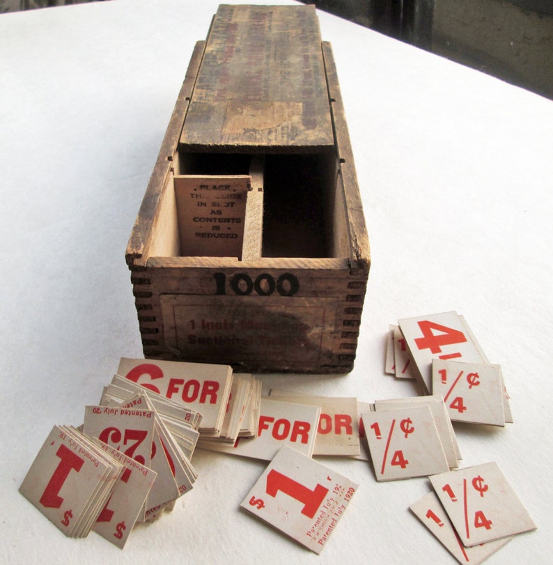 May include: A vintage wooden box with the number "1000" printed on the front, containing small square cards with red numbers and text. The cards read "1", "4", "1/4", and "FOR". The box has a sliding lid and a slot inside.