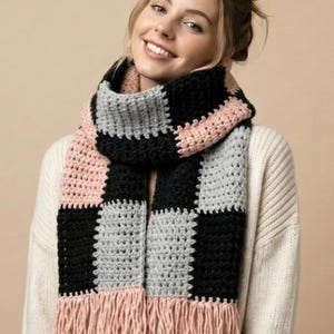 May include: A handmade crochet scarf in a checkerboard pattern of black, gray, and pink. The scarf has a fringe of pink yarn at the bottom. The scarf is worn over a white sweater.