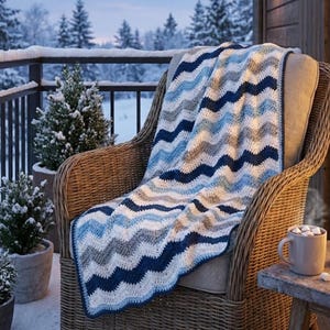 May include: A blue, white, and gray chevron-patterned crochet blanket draped over a wicker chair. The blanket is on a snowy porch with evergreen trees in the background. A mug of hot chocolate sits on a small wooden table.