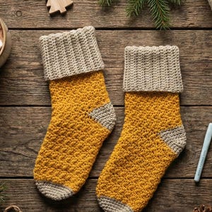 May include: Two handmade, yellow and grey crochet Christmas stockings. The stockings have a textured stitch pattern and a folded-over cuff. They are displayed on a rustic wooden surface, with festive decorations in the background.