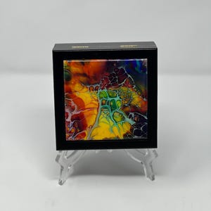 Pode incluir: Uma pequena obra de arte emoldurada em preto com um design abstrato vibrante. A obra apresenta uma mistura colorida de tons amarelos, vermelhos, verdes e azuis, criando uma peça dinâmica. A moldura é quadrada e assenta num expositor transparente.