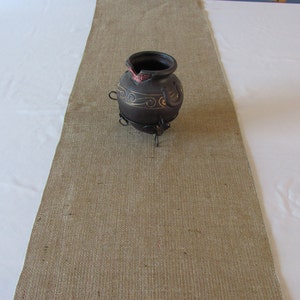 May include: A brown ceramic pot with gold accents sits on a metal stand on a brown burlap table runner.