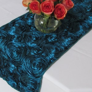 May include: Teal blue satin table runner with a rosette design. The runner is decorated with a vase of orange roses.
