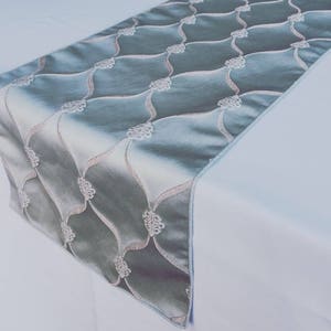 May include: A light blue table runner with a white embroidered pattern. The pattern features a repeating design of curved lines and floral motifs.