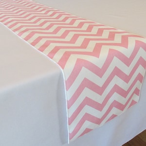 May include: Pink and white chevron table runner. The runner is made of a soft, smooth fabric and has a modern, geometric design.