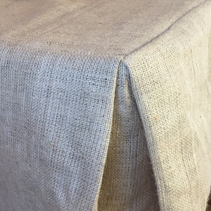 May include: A natural burlap table runner with a simple, folded edge. The runner is made of a coarse, woven fabric with a natural, earthy color.
