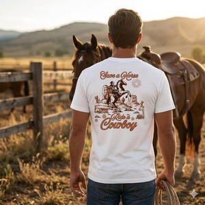 Könnte beinhalten: Weißes T-Shirt mit einem Western-Motiv auf dem Rücken. Die Grafik zeigt einen Cowboy auf einem Pferd, Kakteen und den Text "Save a Horse Ride a Cowboy" in Braun und Orange. Die Person trägt eine blaue Jeans und hält ein Seil.