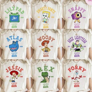 May include: Nine off-white t-shirts featuring various Toy Story character designs. Each shirt has a character name in a bold font, the Toy Story logo, and the year of the character's debut. Characters include Lilypad, Smarty Pants, Snappy, Atlas, Woody, Buzz Lightyear, Jessie, Rex, and Forky.