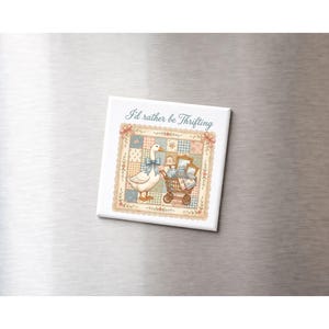 Peut inclure: Aimant carré avec une illustration fantaisiste. Il présente une oie blanche portant un nœud bleu, un chariot rempli d'articles et le texte "I'd rather be Thrifting" dans une police décorative. Le fond a un motif patchwork.